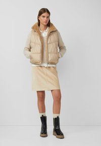 Beige gepolsterte Weste mit Fellkragen, beiger Strickpullover, cremefarbenes Hemd, beiger Rock, schwarze Stiefeletten mit dicken Sohlen, helle Socken.