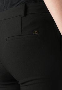 Crne, krojene hlače s suptilnom teksturom i malim zlatnim logotipom na stražnjem džepu, uz klasičnu dizajn džepova.