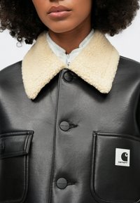 Czarna skórzana kurtka z beżowym futrzanym kołnierzem, z dwoma przednimi kieszeniami i czarnymi guzikami. Mała biała metka z logo na lewej kieszeni.