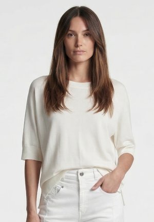 Woman with long brown hair wearing a loose white top and white pants, one hand in pocket, neutral expression, plain light background.