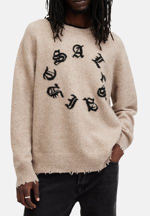 Maglione a maglia beige con un design del logo nero rotondo. Presenta un collo a giro, maniche lunghe e un orlo sfilacciato. Texture morbida, vestibilità casual.