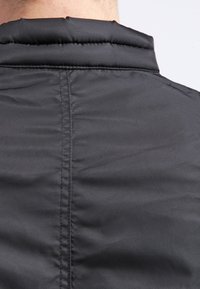 Svart puffjacka med hög krage, slät textur och synliga sömmar på baksidan. Materialet verkar glänsande och vattenavvisande.