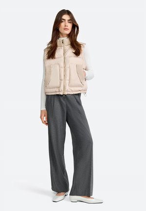 Beige gewatteerd vest met een hoge kraag, rits aan de voorkant en geribbelde zakaccenten. Gecombineerd met grijze wijde broek en witte platte schoenen.