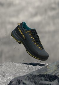 Μαύρο παπούτσι πεζοπορίας με γκρι λεπτομέρειες, κίτρινα κορδόνια και τυρκουάζ εσωτερικό. Διαθέτει υφή στην επάνω επιφάνεια και ανθεκτική σόλα, σχεδιασμένα για υπαίθρια δραστηριότητα.