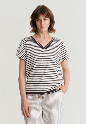 Femme portant un haut à rayures noires et blanches avec col en V et manches courtes, et un pantalon à rayures beige et blanc, debout avec une main dans la poche.