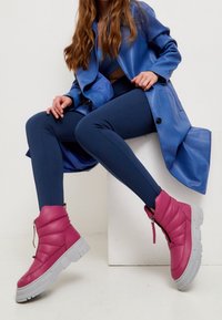 Botas acolchadas rosas hasta el tobillo con suela gris gruesa, combinadas con leggings azul marino y un abrigo de cuero azul. Se destaca la textura suave y los detalles de cierre.