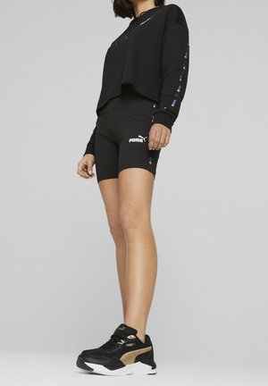Personne portant un short noir Puma, un sweat-shirt court noir avec des accents de couleur sur les manches, et des baskets Puma noires et dorées.
