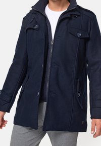 Man wearing a partially zipped navy blue coat over a white shirt and light grey pants against a plain background.