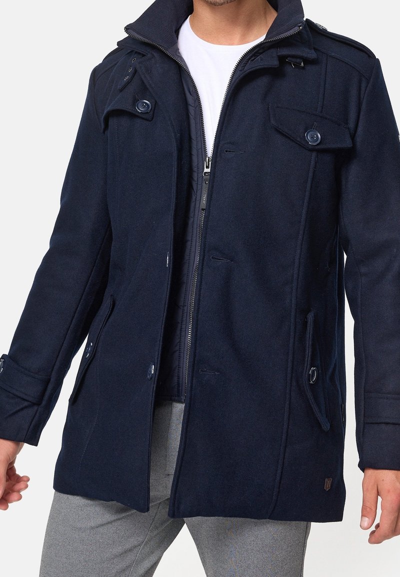 Man wearing a partially zipped navy blue coat over a white shirt and light grey pants against a plain background.