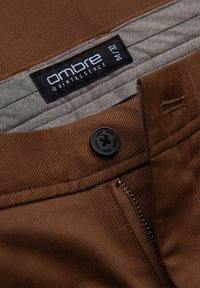 Braune Stoffhose mit strukturiertem Gewebe, ausgestattet mit einem schwarzen Knopf, einem Reißverschluss und einem grauen Innenfutter mit einem Etikett, das "ombre" markiert ist.