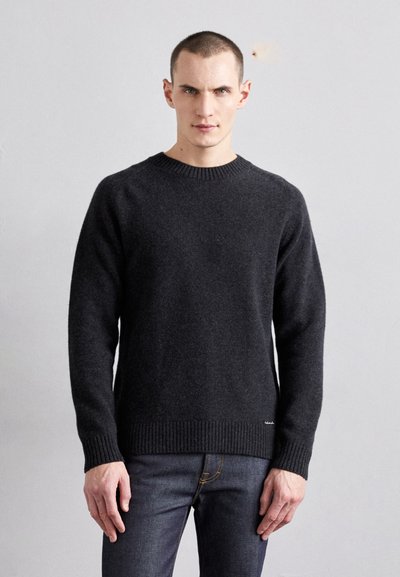 Dunkelgrauer Wollpullover mit Rundhalsausschnitt, gerippten Bündchen und Saum. Getragen mit blauen Jeans, die ein sichtbares Markenetikett an der Seite haben.