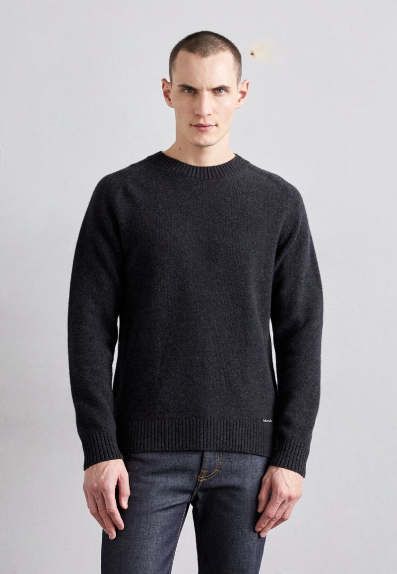 Mørkegrå ullgenser med rund hals, ribbede mansjetter og hem. Brukt med blå jeans, som har en synlig merkeetikett på siden.