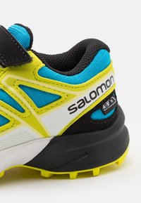 Sport shoe with a black and yellow rubber sole. Upper is blue and yellow mesh with black accents. Features a Velcro strap and waterproof label.