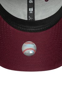 Bordeauxrotes Basecap mit schwarzem Schirm, auf dessen Unterseite sich ein runder Aufkleber befindet, der einem Baseball ähnelt; strukturierte Stoffoberfläche.