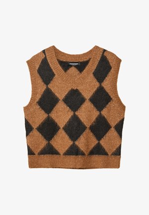 Personne portant un gilet en maille à losanges marron et noir sur une chemise blanche, un jean large sombre avec ourlets retroussés et des chaussures foncées sur un fond neutre.