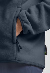Giacca in pile blu scuro con tasca laterale con zip, realizzata con una texture morbida e rifinita con bordi neri a contrasto. Presenta un'etichetta Polartec.