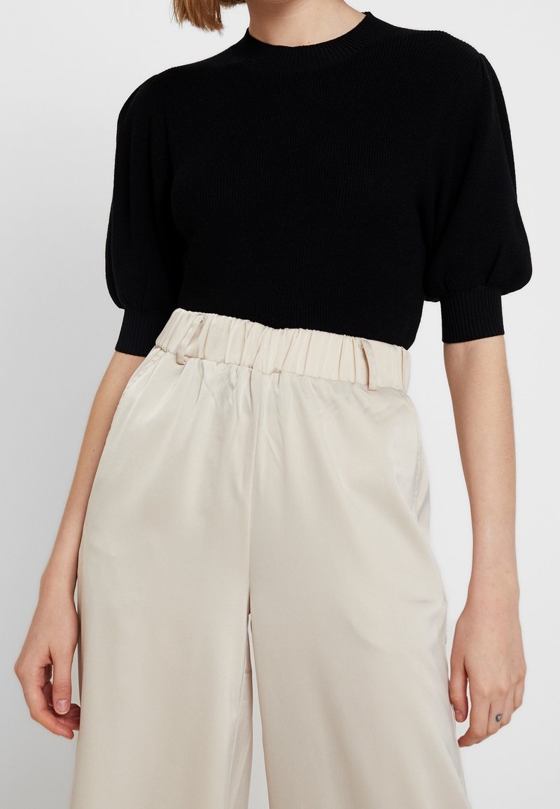 Černý pletený svetr s krátkými nafouknutými rukávy, spárovaný s krémovými hedvábnými kalhotami s vysokým pasem a širokými nohavicemi, které vystihují hladkou texturu.