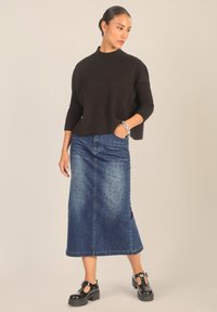 Zwarte gebreide trui met geribbelde mouwen, gecombineerd met een lange spijkerrok met stippenversieringen. Zwarte plateau-schoenen maken de look compleet.
