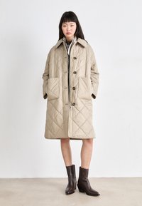 Manteau beige matelassé avec de grandes poches, un devant à boutons et un large col. Associé à des bottes à talons marron, le tout sur un fond clair.