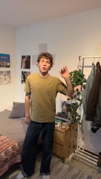 Noor inimene kollast särki ja tumedaid pükse kandes seisab magamistoas, lehvitab käega voodi, taime ja riidepuu lähedal.