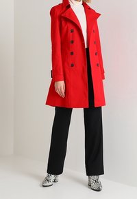 Červený trench coat s dvouřadým designem, černými knoflíky a klopovým límcem. V kombinaci s černými kalhotami a vzorovanými botami.