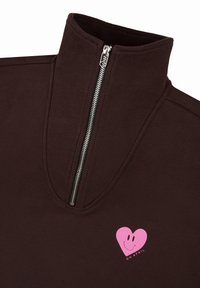 Felpa marrone con zip caratterizzata da un colletto alto, texture a coste e un design di un cuore rosa con un faccino sorridente sul petto.