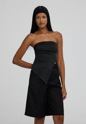 Top nero senza spalline realizzato in tessuto liscio, con orlo diagonale e dettagli arricciati. Abbinato a pantaloni corti neri larghi per un look coeso.