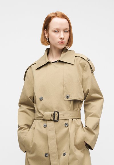 Νεαρός με κοντά κόκκινα μαλλιά που φοράει ένα μπεζ παλτό τύπου trench με τα χέρια στις τσέπες, ποζάροντ ας με φόντο ένα απλό λευκό φόντο.