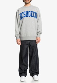 Sudadera gris con letras azules "DCSHOECO", combinada con pantalones de mezclilla oscuros y zapatillas blancas. Corte relajado, con puños y cintura acanalados.