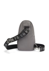 Graue Stoff-Slingtasche mit runder Form. Mit einem schwarzen Tragegurt mit „SAND“-Schriftzug und silbernem Zubehör. Strukturierte Oberfläche.