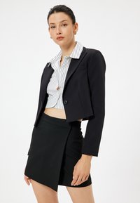 Schwarze, taillierte Blazerjacke mit einem einzigen Knopf, figurbetontem Design und langen Ärmeln, kombiniert mit einem schwarzen Wickelrock, der einen minimalistischen Stil betont.