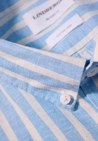 Camicia in cotone e lino a righe blu chiaro e bianche, con colletto, caratterizzata da bottoni bianchi e etichette visibili che mostrano informazioni sul marchio e sul materiale.