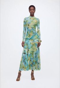 Γυναίκα με μακρυμάνικο φόρεμα με λουλουδάτο σχέδιο σε μπλε και πράσινους τόνους, φορώντας χρυσά σανδάλια και βραχιόλια, που στέκεται μπροστά σε λευκό, απλό φόντο.