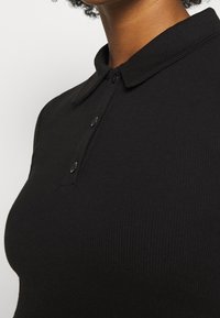 Schwarzes geripptes Poloshirt mit Kragen, ausgestattet mit zwei Knöpfen und einem strukturierten Stoff. Nahaufnahme, die die Passform rund um den Hals und die Schulter zeigt.