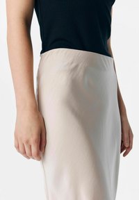 Lichtroze, gladde stoffen rok met een aangesloten tailleband, gecombineerd met een zwart geribbelde top. Gezien van de taille naar beneden, met de hand rustend op de rok.