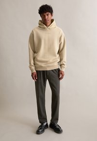 Beżowa bluza z kapturem o luźnym kroju i ściągaczach na rękawach, zestawiona z ciemnoszarymi spodniami i czarnymi butami. Prosty fason, bez widocznych wzorów.