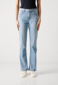 Jeans de mezclilla de tiro alto, de campana y color azul claro, con cierre de botón y cinco bolsillos, combinados con zapatillas blancas, sobre un fondo neutro.