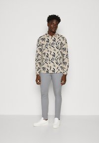 Langermet skjorte med knapper i beige stoff med svarte bladmønstre, kombinert med smale grå bukser og hvite sneakers.