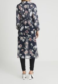 Žena oblečená v průhledných námořnických květinových šatech přes černé kalhoty a bílé vysoké podpatky, stojící zády k jednoduchému pozadí.