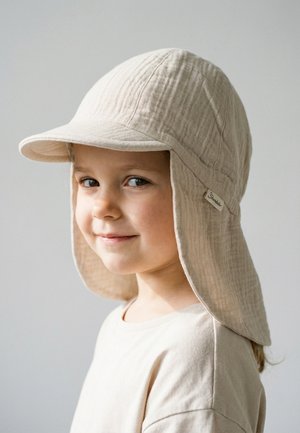 Junges Kind mit einem hellbeigen Sonnenhut mit Nackenschutz und einem passenden hellbeigen Hemd, leicht lächelnd, vor einem einfarbigen Hintergrund.