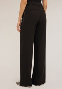 Wijdvallende zwarte broek van gladde stof, met een hoge taille, twee achterzakken en subtiele zijkanten. Textuur schoenen zijn zichtbaar.