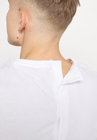 Camiseta blanca con cierre de botón en la parte posterior del escote. Tejido suave y ligero con una textura suave.