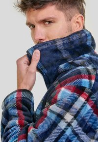 Hombre levantando el cuello de una chaqueta de forro polar a cuadros azul, rojo y blanco, cubriendo parcialmente su rostro, con un fondo neutro.