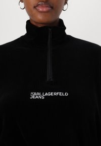 Black zip-up pullover made of ribbed fabric; features "KARL LAGERFELD JEANS" in white embroidery on the front.