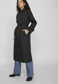 Trench coat negru cu talie accentuată prin curea, butoane duble și buzunare laterale. Purtați-l cu blugi evazați albaștri și bocanci negri până la gleznă.