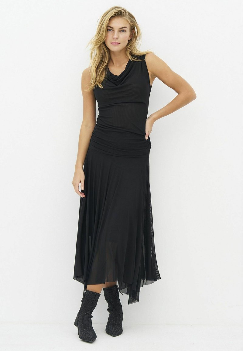 Femme debout dans une robe noire sans manches et des bottines noires, posant avec une main sur la hanche devant un fond blanc uni.