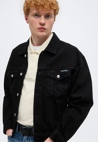 Νεαρός με σγουρά κόκκινα μαλλιά, φορώντας μαύρο τζιν μπουφάν πάνω από μπεζ πόλο και μπλε τζιν, στέκεται μπροστά σε λευκό, απλό φόντο.