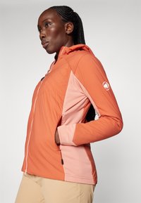 Lightweight outdoor jacket in coral with a hood, zip closure, and pink side panels. Features pockets and textured detailing on the fabric.