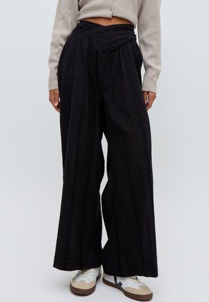 Frau trägt hoch taillierte, weiter geschnittene schwarze Hosen mit dezenten vertikalen Mustern und beigen Turnschuhen, kombiniert mit einer hellen, kurzen Strickjacke.