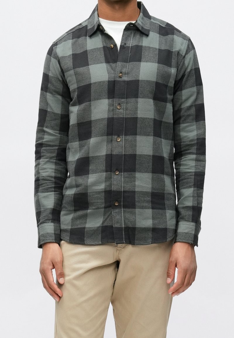 Homme portant une chemise à carreaux noire et grise à manches longues boutonnée sur un sous-vêtement blanc avec un pantalon beige, debout devant un fond uni.